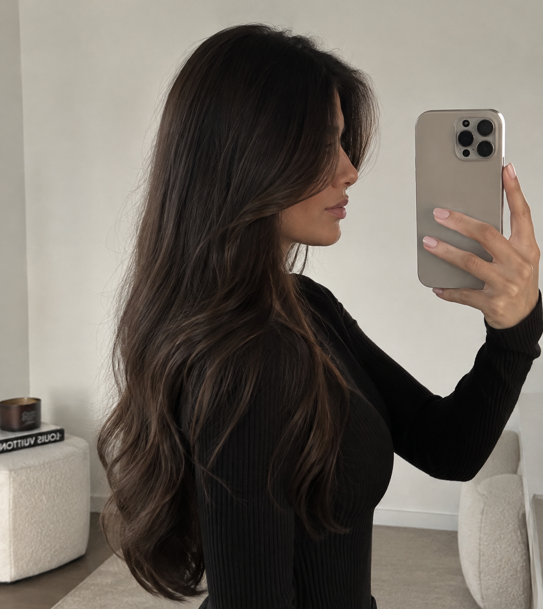 A woman with long, wavy dark brown hair takes a mirror selfie wearing a black ribbed long-sleeve top. She is holding a light beige smartphone in a minimalist indoor setting with a light gray wall and neutral-colored furniture.