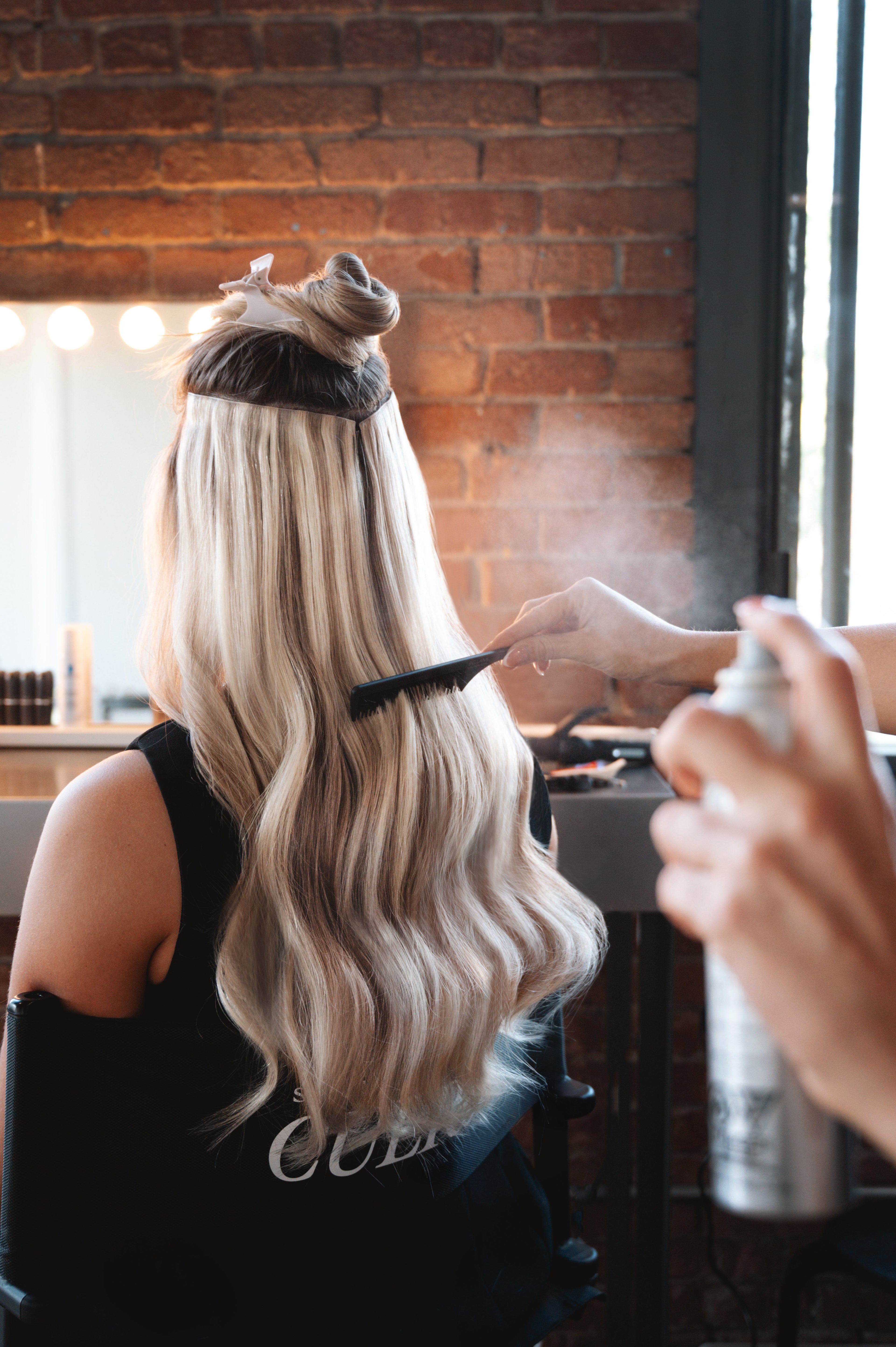 model wearing invisible clip in 2 rows in Russian Nova, getting her hair brushed and sprayed in a hair salon in Montreal / Mannequin portant deux rangées d’extensions à clips invisibles, nuance Russian Nova, se faisant coiffer et vaporiser dans un salon de coiffure à Montréal.